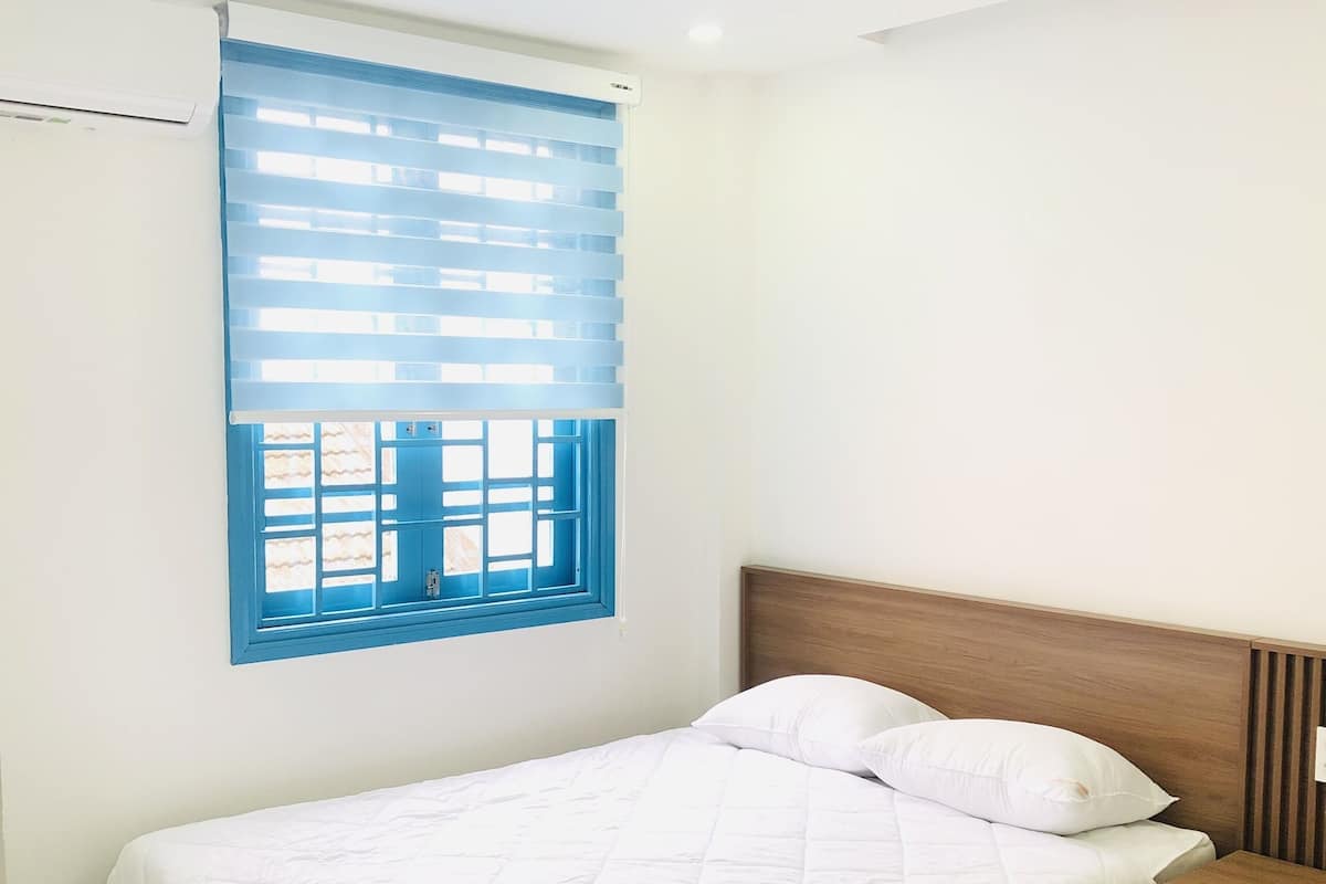 A single bed with a white quilted bedspread, a wooden headboard, and a nightstand with a drawer. A window with blue blinds and a view of the outside is visible.