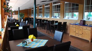 A restaurant with multiple tables and chairs, a wooden cabinet, and a large window with curtains. - Hotel Dominika  (Bratislava)