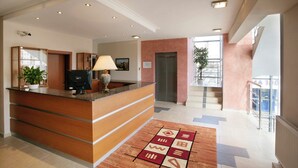 A hotel reception area with a wooden reception desk, a potted plant, a lamp, and a patterned rug. - Hotel Dominika  (Bratislava)