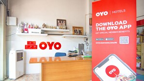 A reception desk with a red OYO Hotels sign, a computer, and a phone displaying the OYO app.