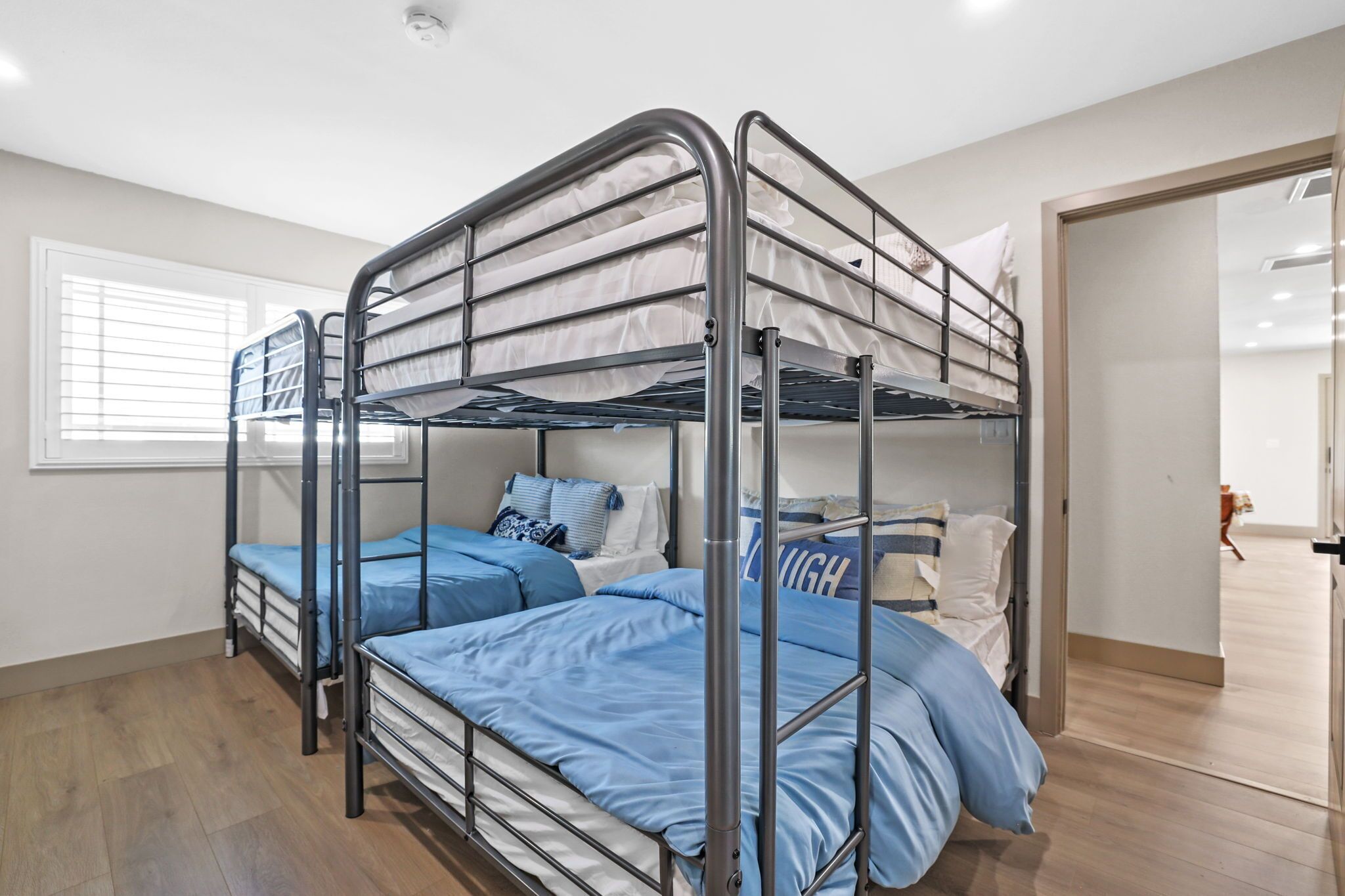 Une chambre avec un lit superposé, une fenêtre, un plancher en bois et une porte donnant sur une autre pièce.