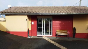 Un bâtiment avec un toit recouvert de tuiles, une façade rouge et jaune, une porte d’entrée en verre et un banc à l’extérieur.