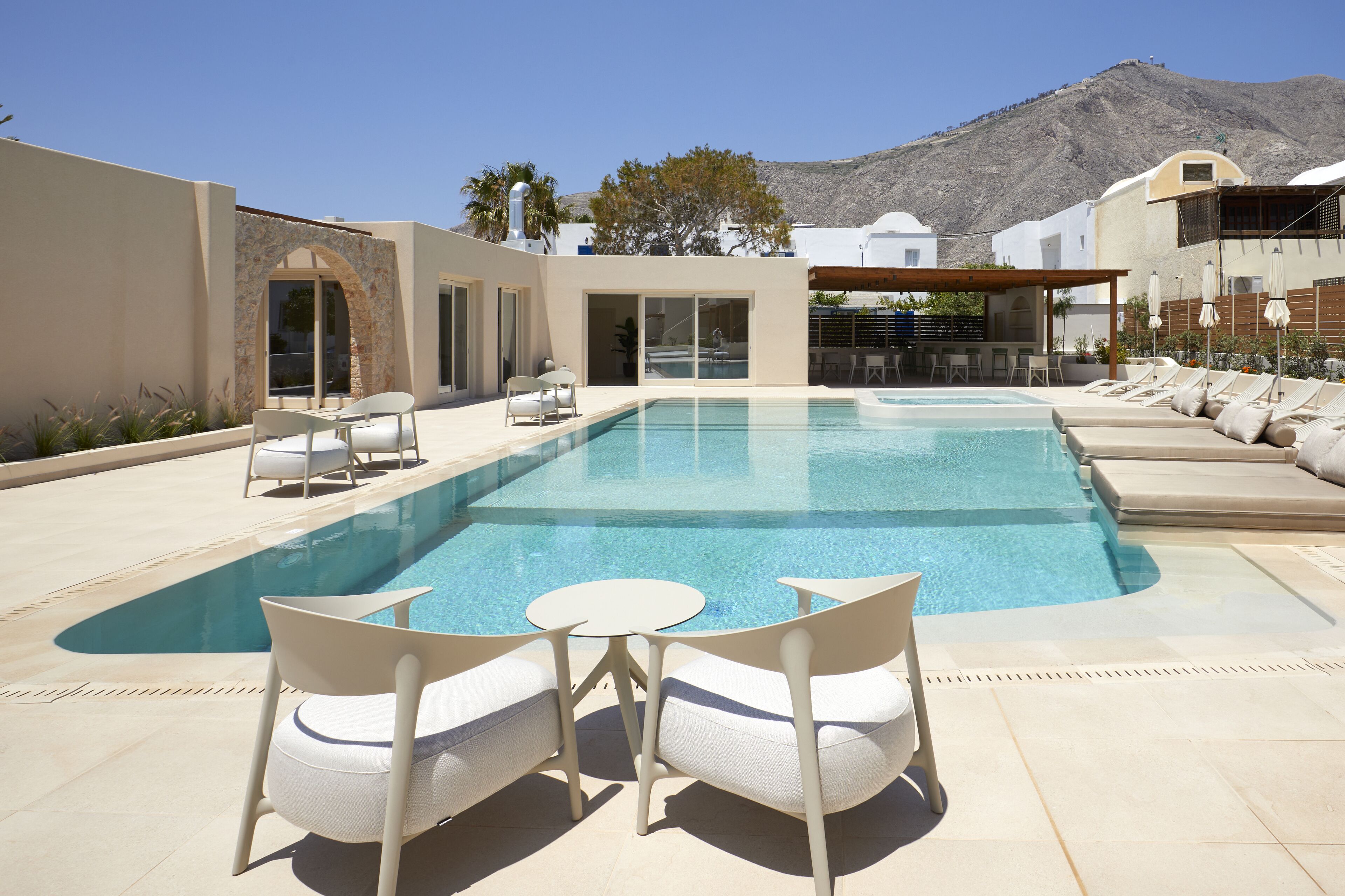 Piscina con sillas de descanso y una mesa, rodeada por un edificio con vistas a la montaña.