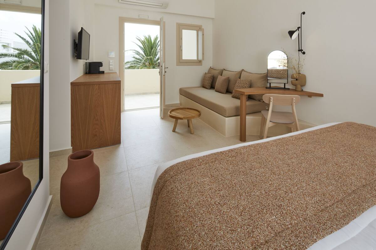A hotel room with a bed, a wooden desk, a sofa, a small table, a chair, a TV, a mirror, and a view of palm trees through a balcony door.