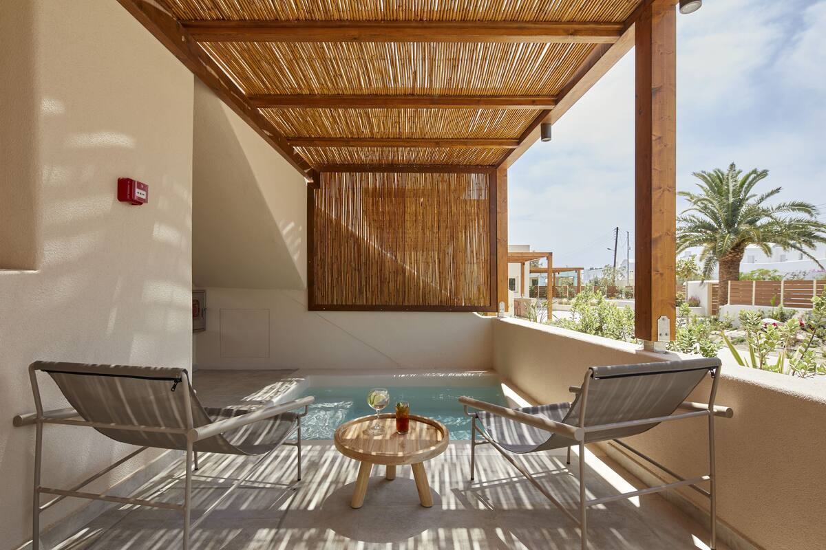 A sunlit outdoor seating area with two chairs and a small round table, a pool, and a view of palm trees and buildings.