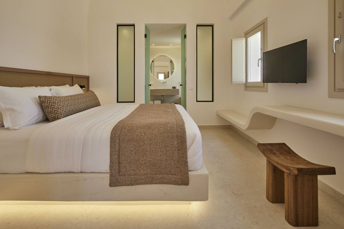 A modern hotel room with a large bed, a wooden bench, a TV mounted on the wall, and a mirror above the sink.