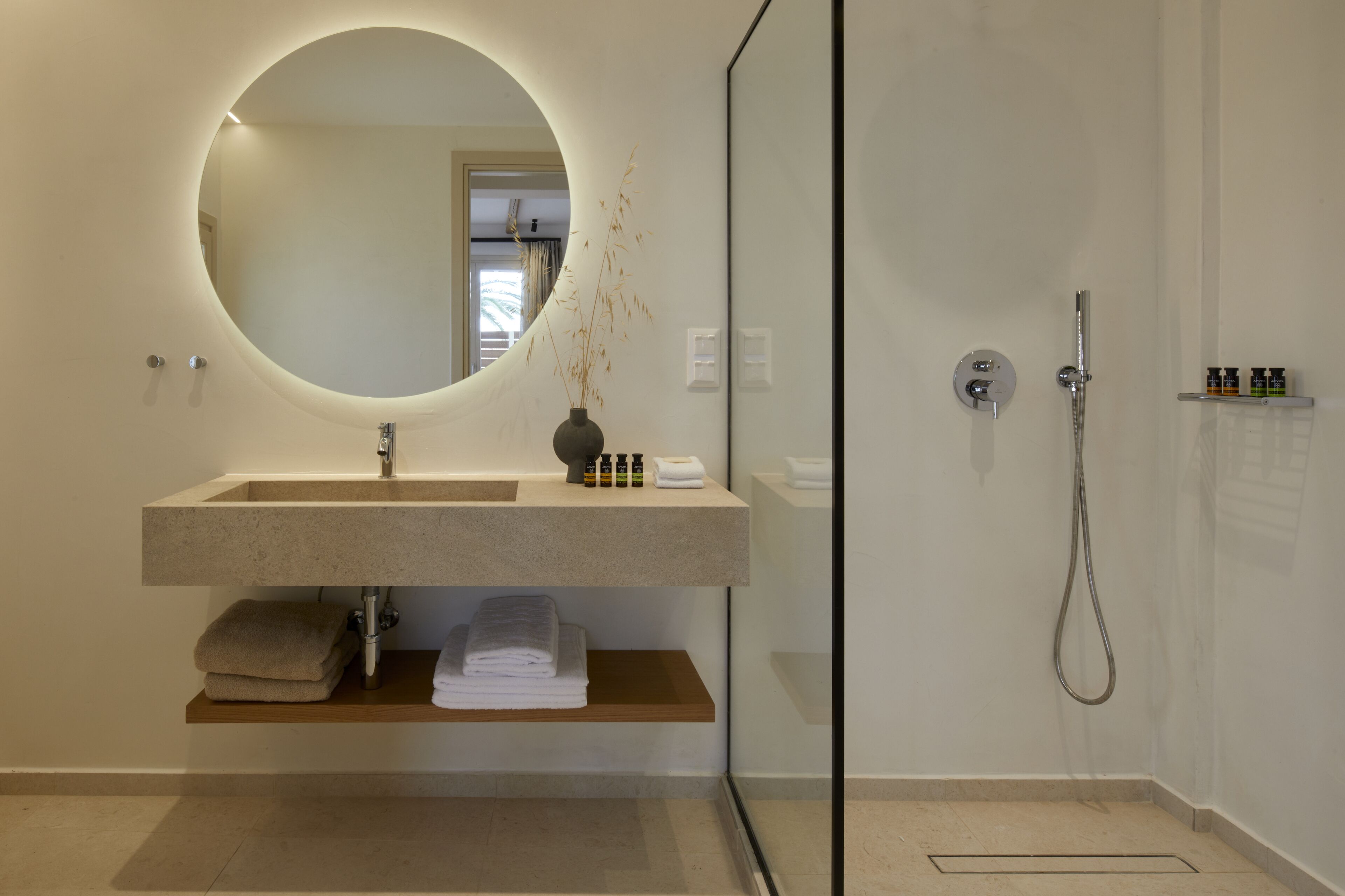 A modern bathroom with a round mirror, a sink, a shower, and a shelf with toiletries.