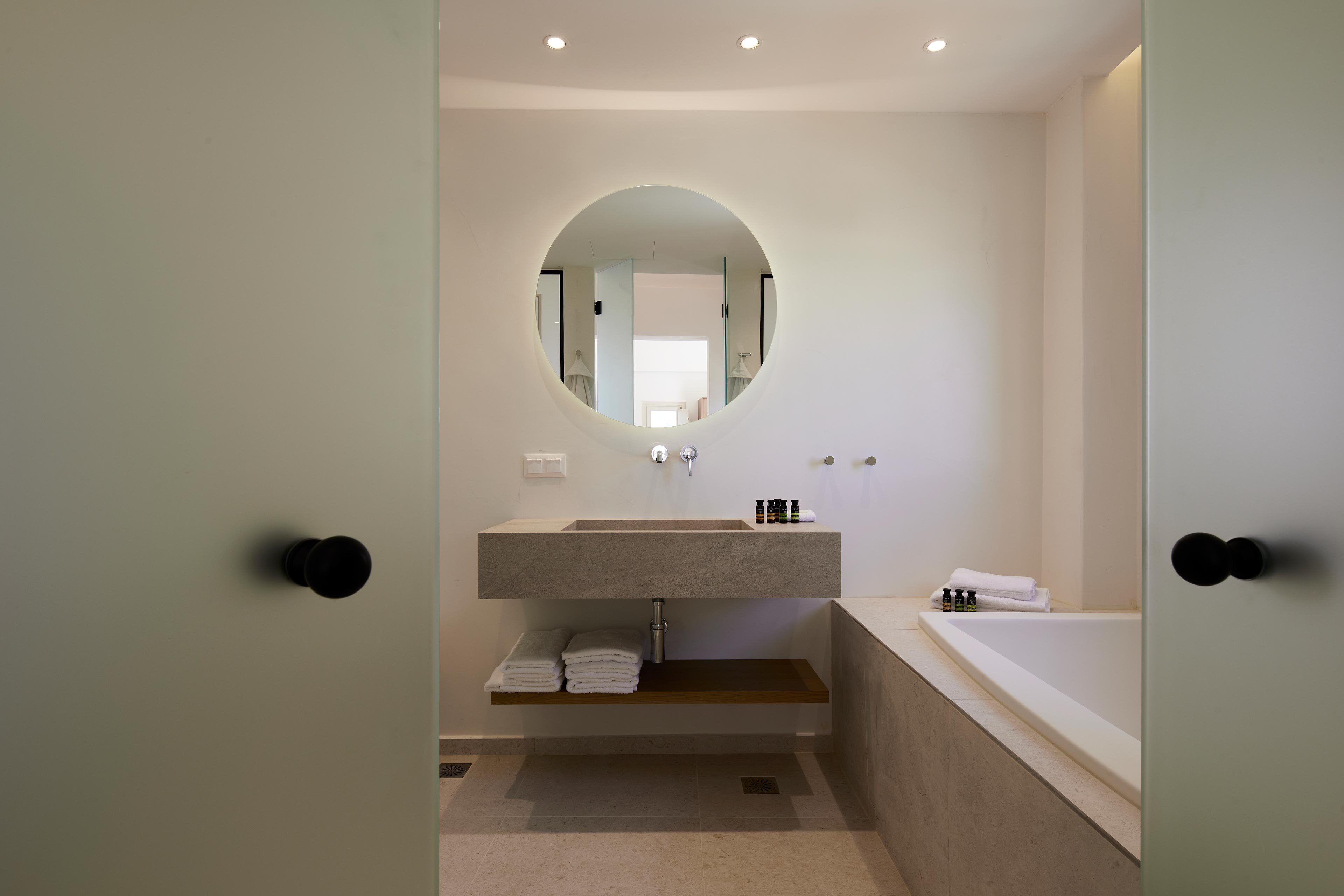 A modern bathroom with a round mirror, a built-in sink, a bathtub, and a shelf with towels.