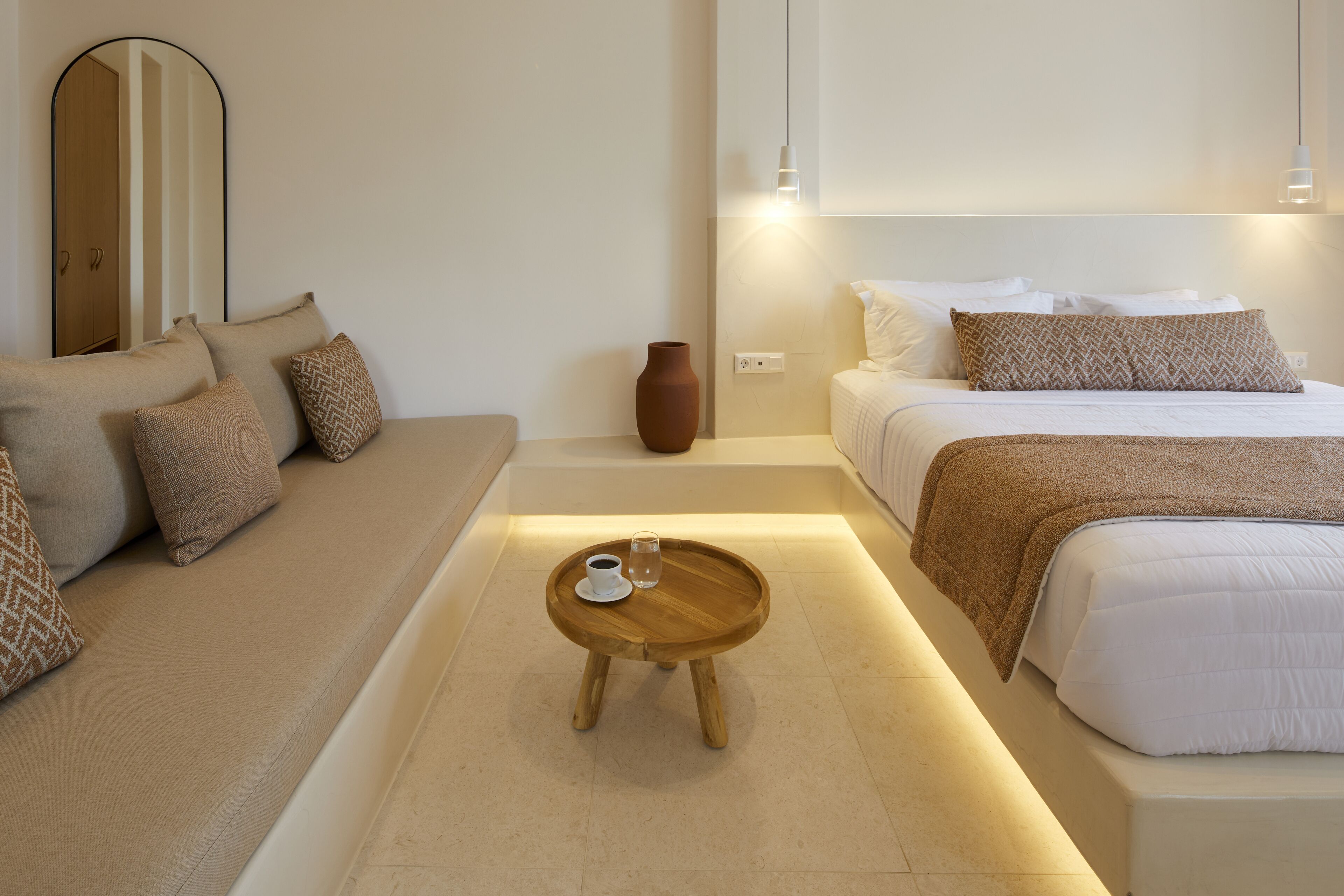 A modern hotel room with a sofa, a small wooden table with a cup and glass, a bed with white bedding, and a vase on the floor.