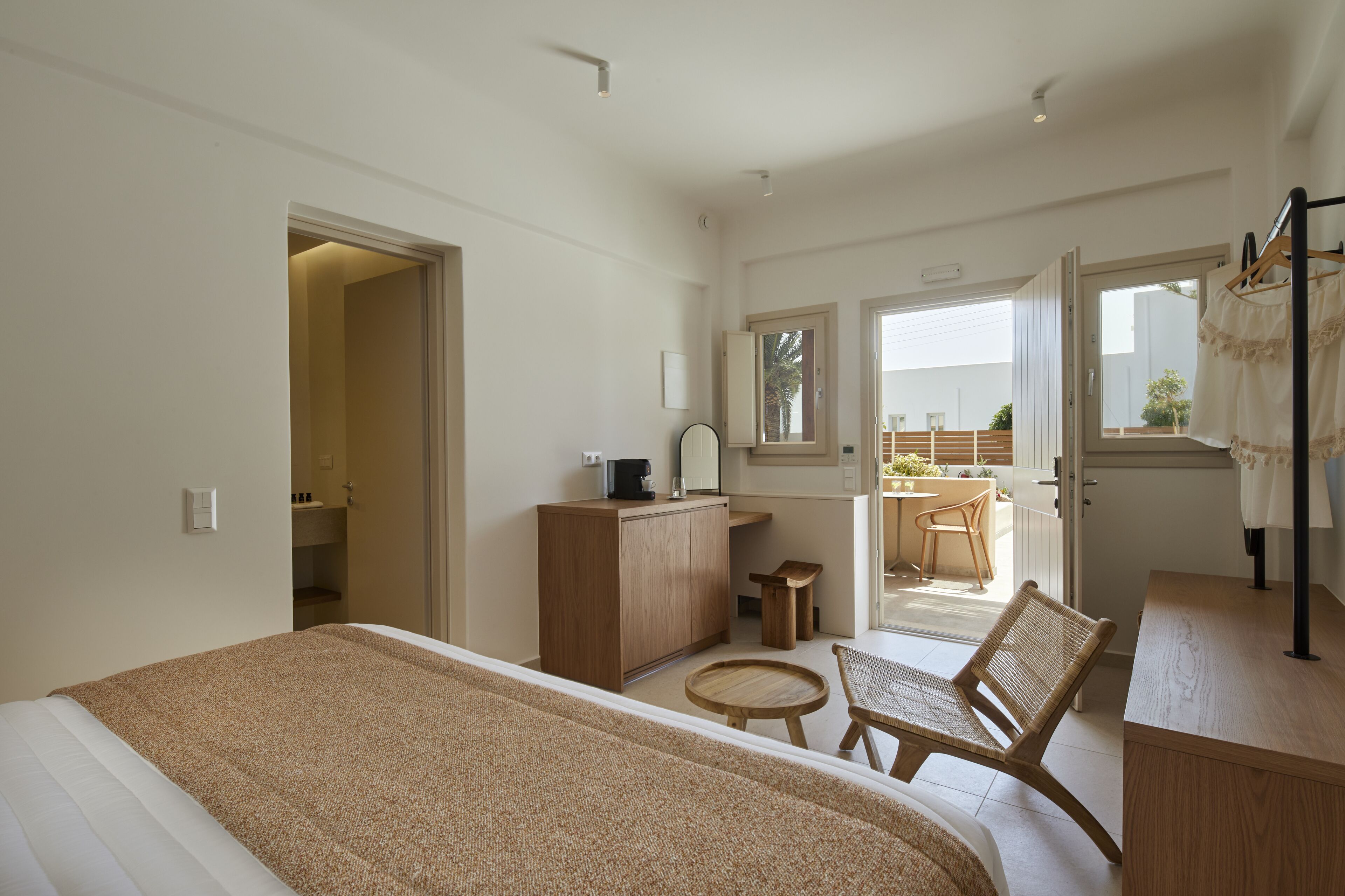 A hotel room with a bed, a small round table, a chair, a wooden dresser, a mirror, and a view of the outside balcony.