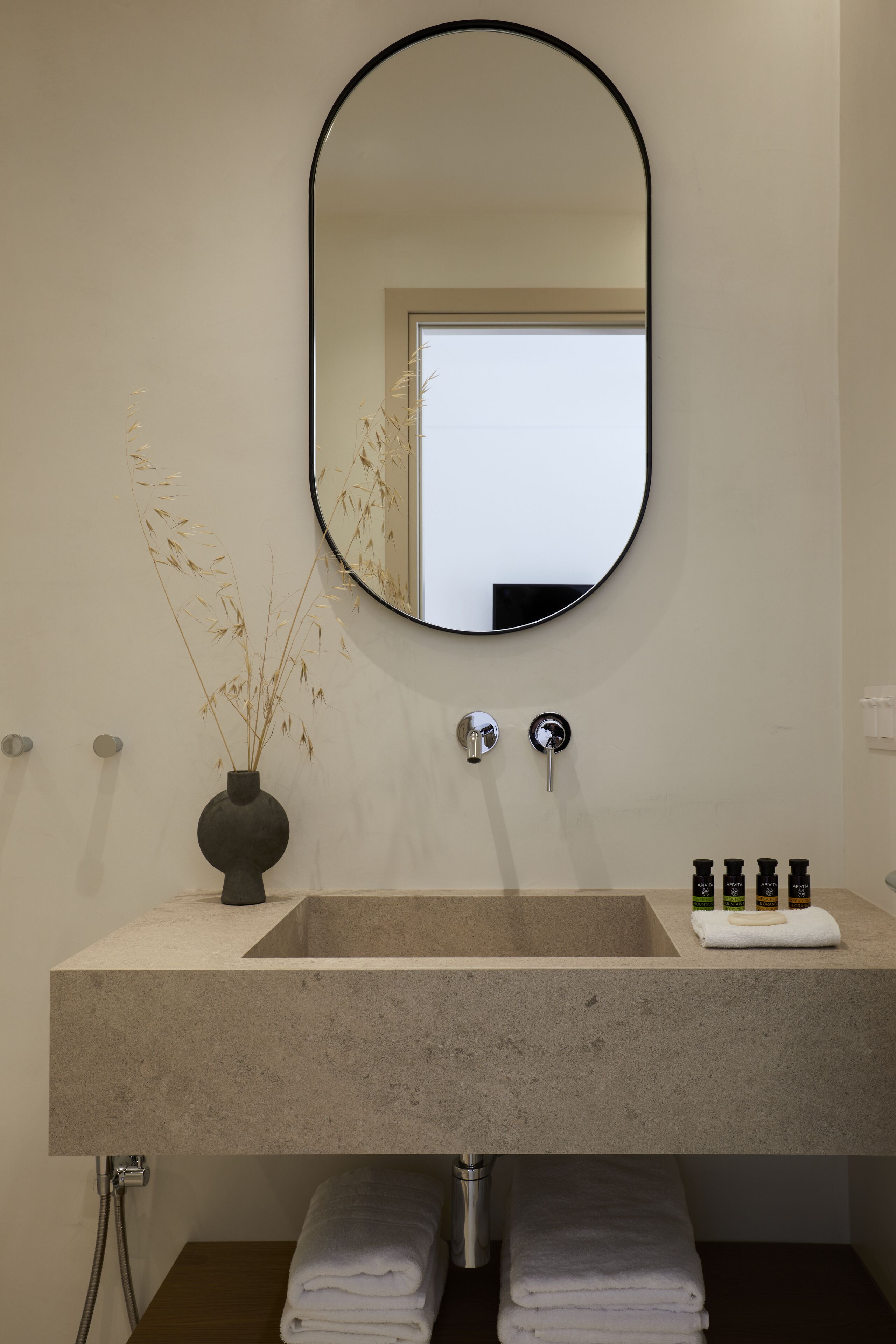 A bathroom with a large oval mirror, a beige sink, a vase with dried plants, and a towel rack.