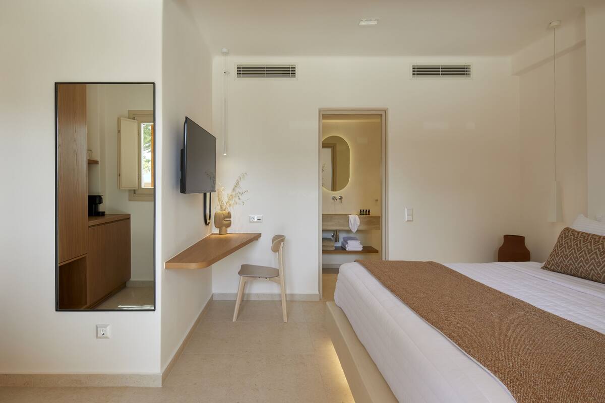 A modern hotel room with a large bed, a desk with a chair, a TV, and a bathroom with a mirror and towel rack.