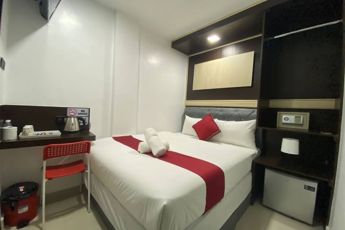 A hotel room with a bed, a desk with a coffee maker, a chair, a trash bin, and a small refrigerator.