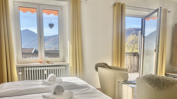 Quarto de hotel com cama, duas janelas com cortinas, radiador e vista para montanhas.