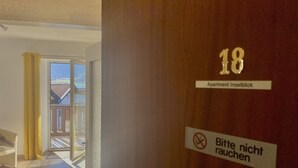 Entrada de um quarto de hotel com uma porta de correr de vidro que dá para uma varanda. Uma placa indica o número do quarto, 18, e um aviso proibindo o fumo.