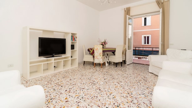 Una sala de estar moderna con un televisor de pantalla plana, una mesa de comedor con sillas, un sofá blanco y un balcón con vistas a un edificio.