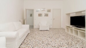 Una sala de estar con un sofá blanco, un sillón blanco, un televisor sobre un soporte blanco y un piso con detalles.