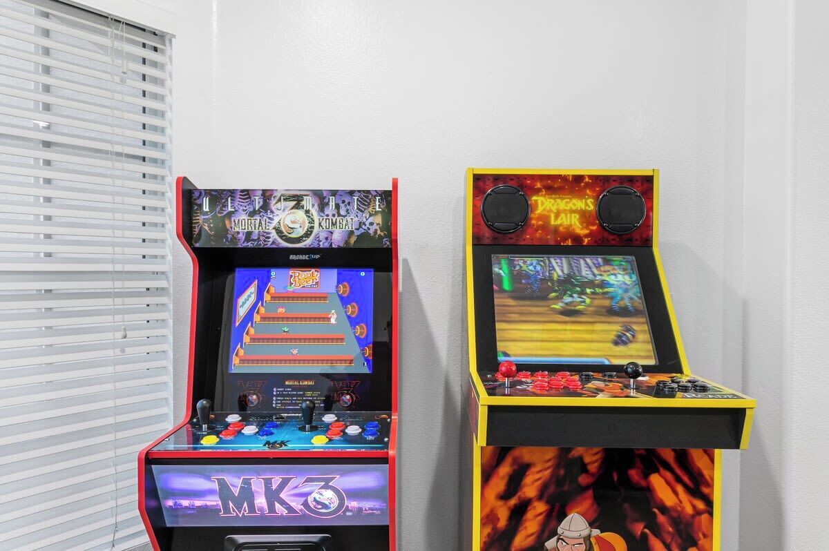Zwei Spielautomaten, einer mit der Aufschrift 'MK3' und der andere mit 'Dragon’s Lair', vor einer weißen Wand mit teilweise sichtbaren Jalousien.