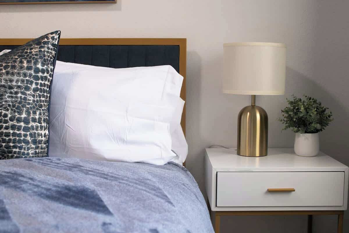 A neatly made bed with white and patterned pillows, a bedside lamp, and a small potted plant on a white nightstand.