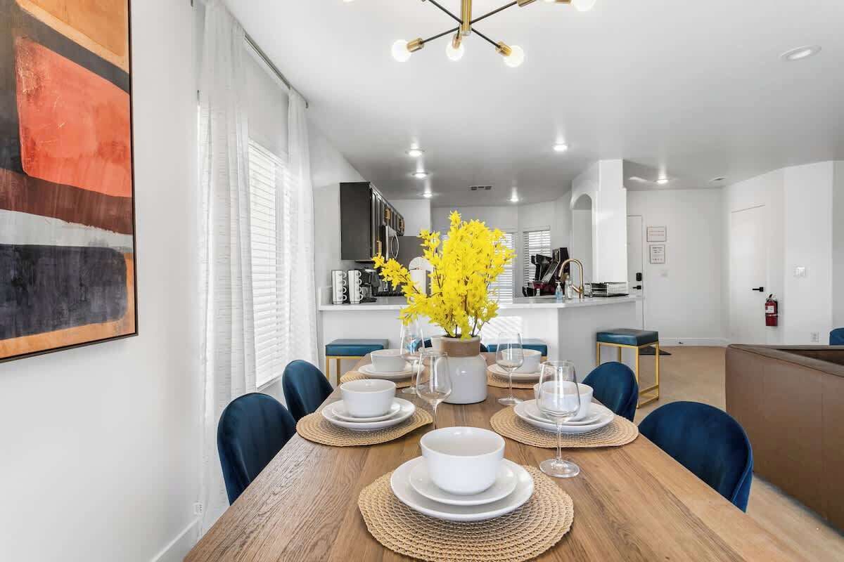 Un'elegante zona pranzo con un tavolo in legno apparecchiato per quattro persone, circondato da sedie blu. Un vaso con fiori gialli è posto al centro.