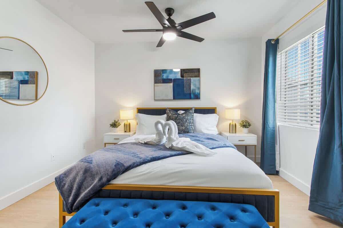 A modern bedroom with a tufted headboard, a ceiling fan, a bed with a blue bedspread and a white pillow arrangement, and a bedside table with lamps.