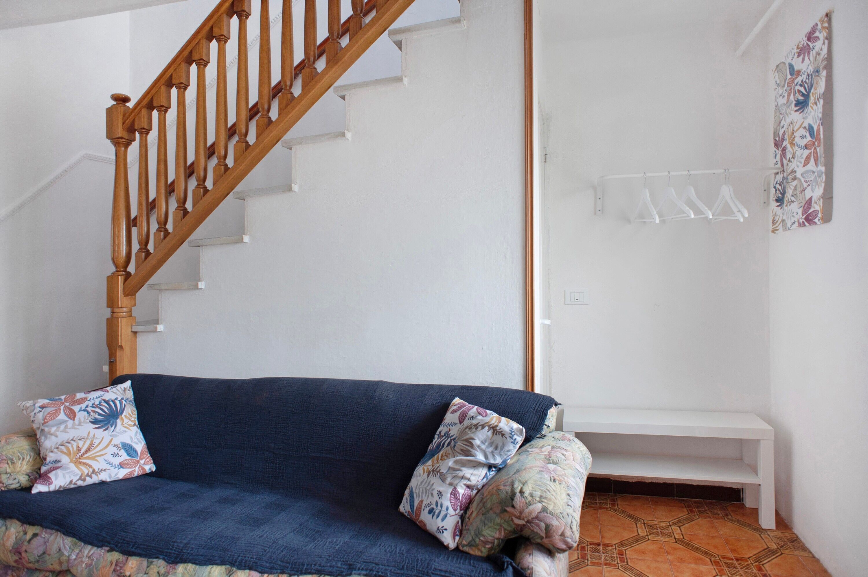 Una habitación con una escalera de madera, un sofá azul con cojines estampados, un banco blanco y un tendedero fijado a la pared.