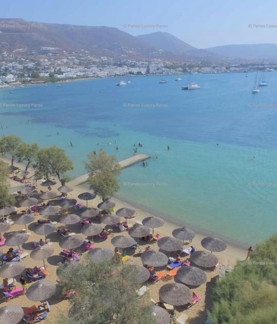 Une vue sur la côte, avec une eau claire, une plage parsemée de parasols et des bateaux à l’horizon.