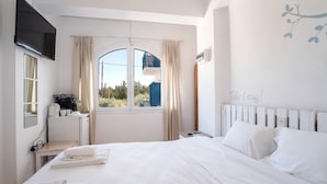 A hotel room with a bed, a desk, a TV, a window with curtains, and a view of the outside. - Studio Apartment 'Livadia Seaside Studios 8' with Shared Terrace, Wi-Fi & Air Conditioning (Paros)
