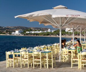 Outdoor dining tables with umbrellas, a beach view, and white buildings in the background. - Studio Apartment 'Livadia Seaside Studios 8' with Shared Terrace, Wi-Fi & Air Conditioning (Paros)