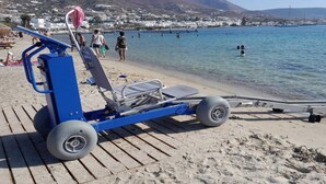 A beachside chair with wheels, a beach umbrella, and a view of the sea and buildings in the background. - Studio Apartment 'Livadia Seaside Studios 12' with Shared Terrace, Wi-Fi and Air Conditioning (Paros)