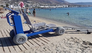 A beachside chair with wheels, a beach umbrella, and a view of the sea and buildings in the background.