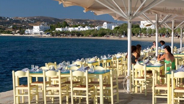 Outdoor dining tables with umbrellas, a beach view, and white buildings in the background.