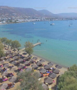A coastal view with clear water, a beach with umbrellas, and boats in the distance.