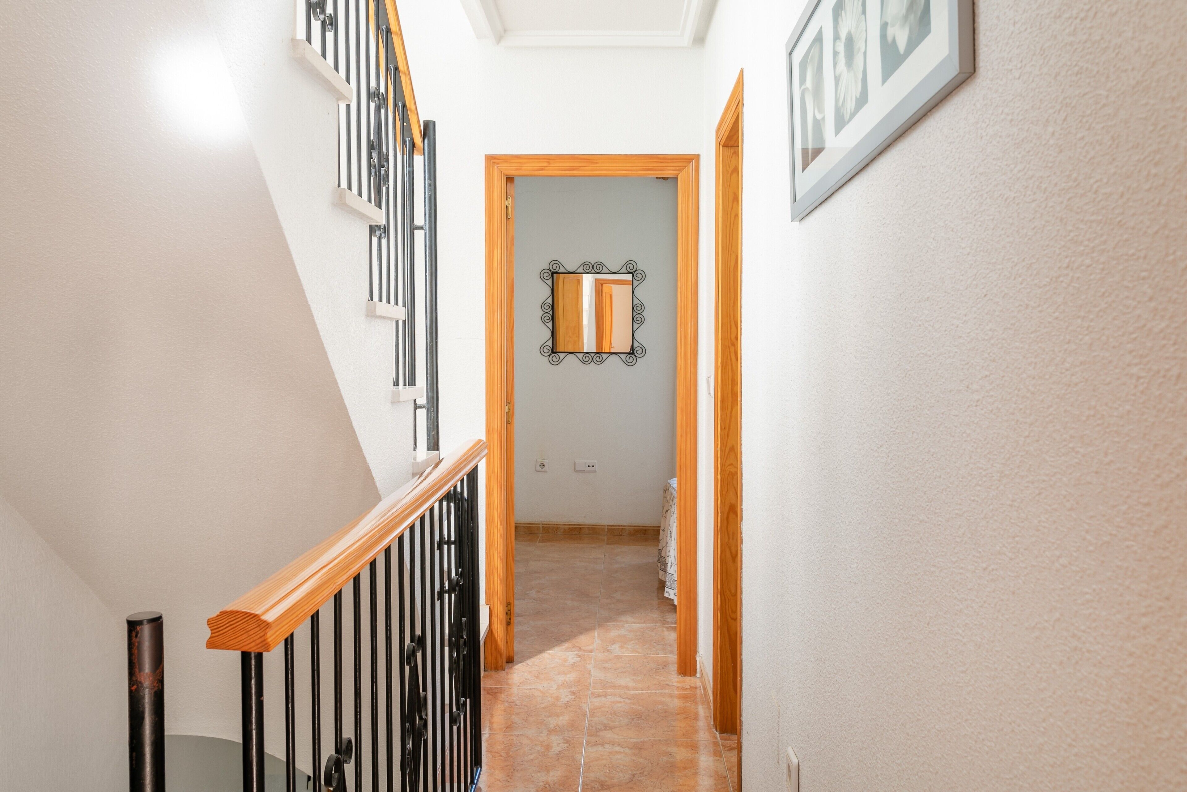 Una escalera con barandas negras, un pasamanos de madera y piso de baldosas que conduce a una habitación con puerta y espejo.