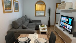 A modern living room with a grey sofa, dining table set with plates and glasses, a TV displaying a show, and framed artwork on the wall. - Bungalow 'Villa Matías' with Sea View, Wi-Fi and Air Conditioning (Gran Alacant)