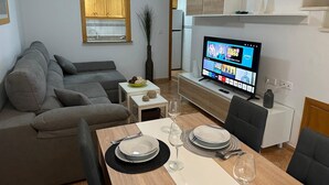 A modern living room with a grey sofa, a dining table set with plates and glasses, a TV displaying a streaming service, and a staircase leading to an upper level. - Bungalow 'Villa Matías' with Sea View, Wi-Fi and Air Conditioning (Gran Alacant)