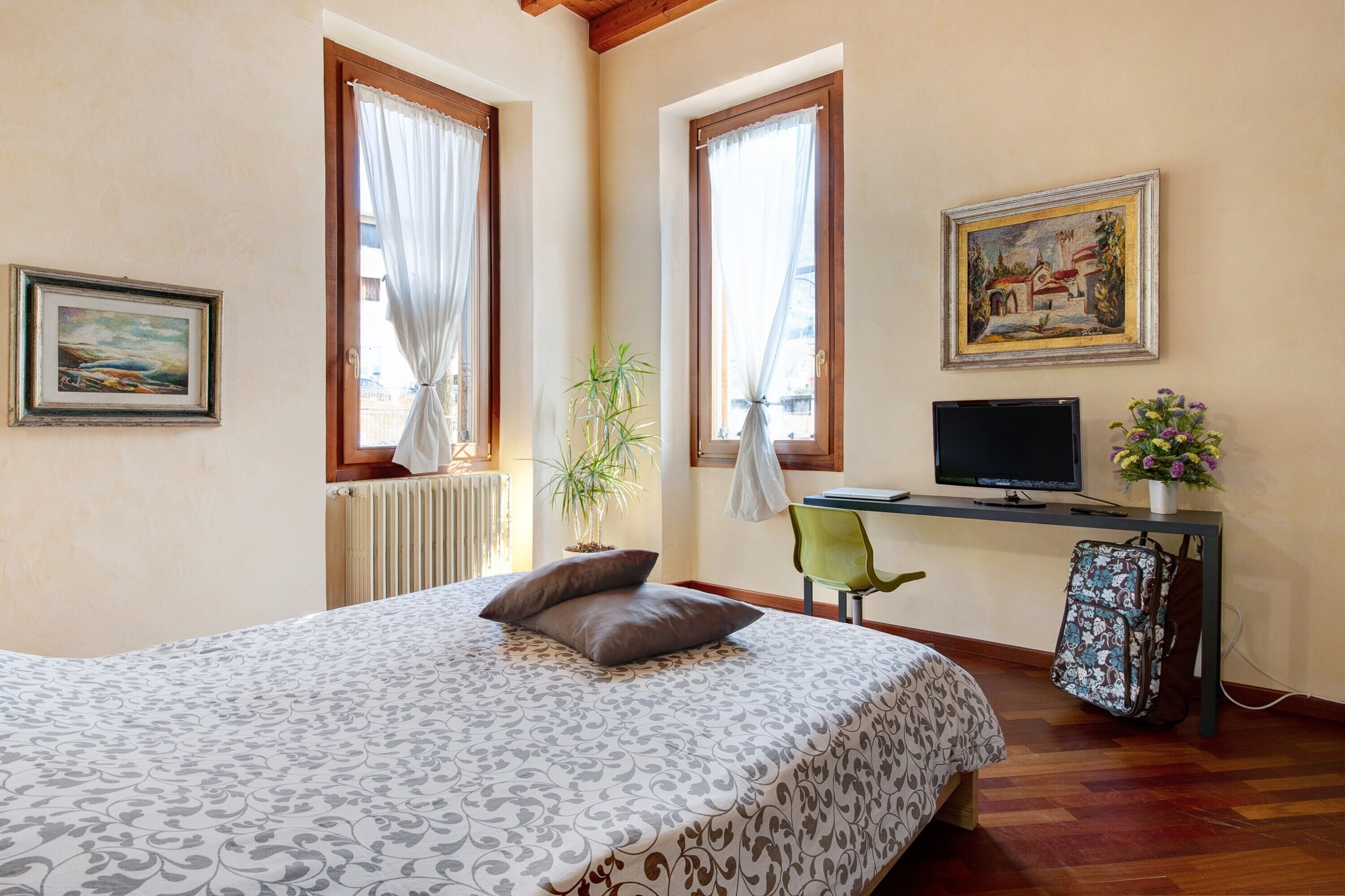 A bedroom with a bed, a desk with a computer, a chair, a radiator, and a window with curtains.
