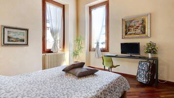 A bedroom with a bed, a desk with a computer, a chair, a radiator, and a window with curtains.