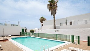 Una piscina con valla verde, sillas de descanso blancas y una palmera.