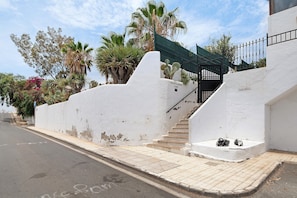 A white building with a staircase leading to a gated entrance, surrounded by palm trees and cacti.