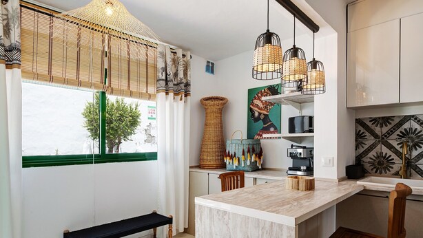 Una cocina con una isla de mármol, sillas de madera, un banco, una cesta de mimbre, una cafetera y una lámpara colgante.