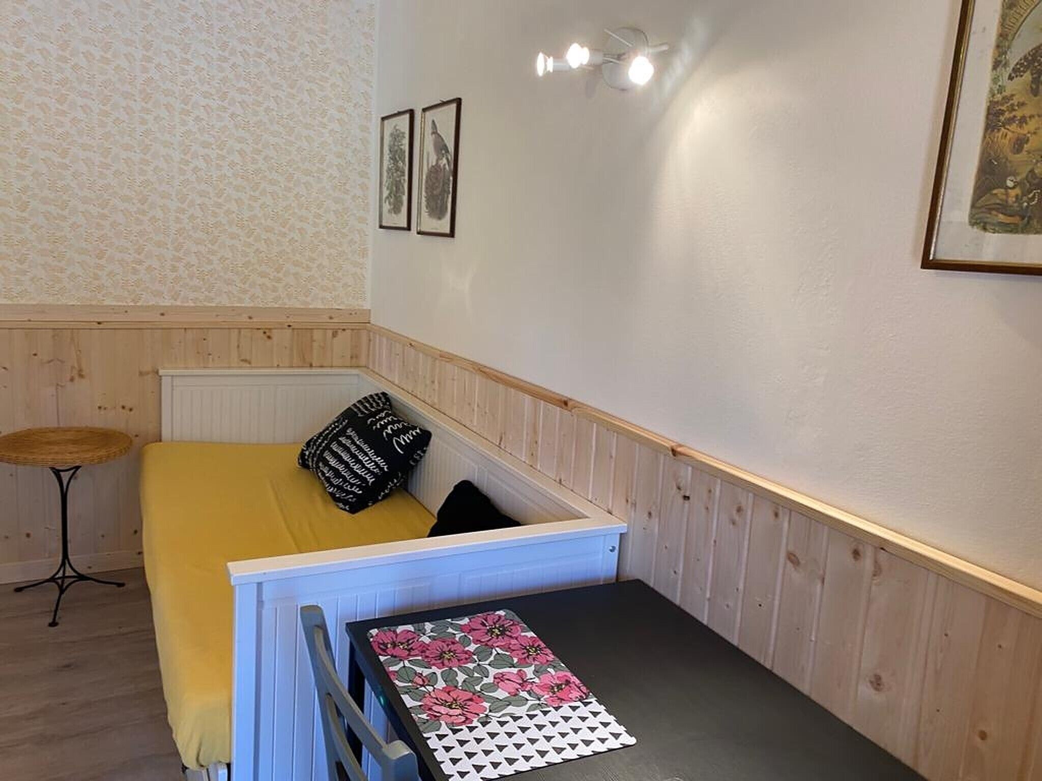 A small room with a bed, a table, a chair, a small stool, and framed pictures on the wall.