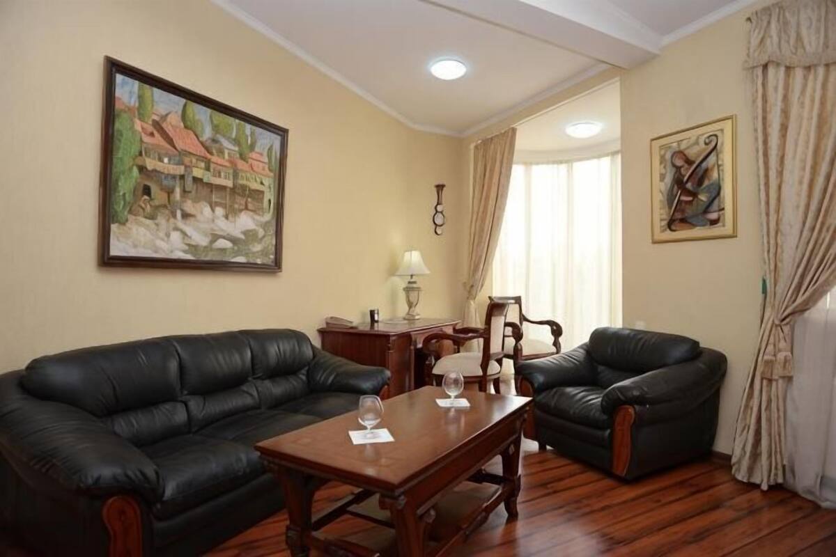 Ein Wohnzimmer mit einer schwarzen Ledersofa, einem Holztisch, einer Lampe und zwei Sesseln. An der Wand hängt ein Gemälde und es gibt ein Fenster mit Vorhängen.