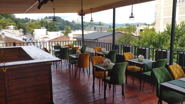 An outdoor dining area with wooden tables and chairs, a wooden bar, and a view of buildings and trees in the background. - Aeetes Palace  (Kutaisi)