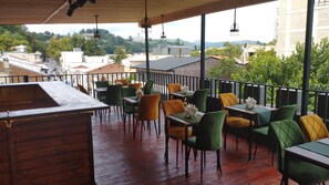 An outdoor dining area with wooden tables and chairs, a wooden bar, and a view of buildings and trees in the background. - Aeetes Palace  (Kutaisi)