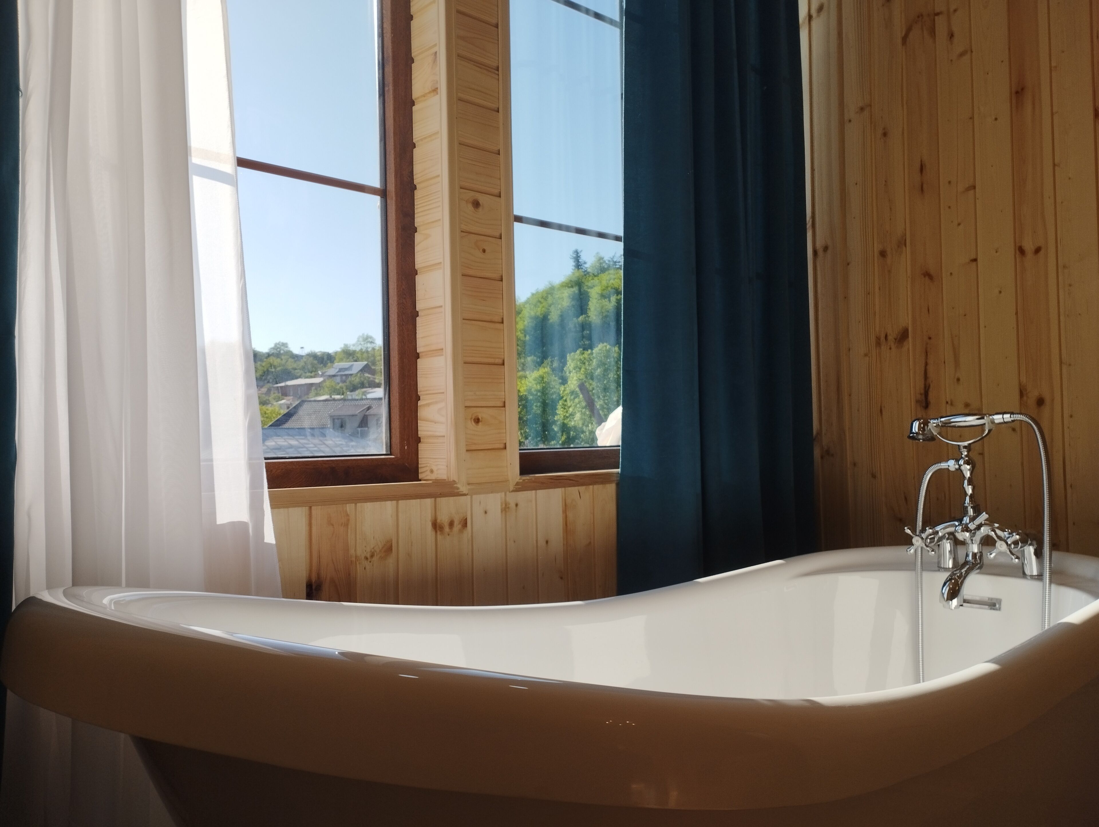 Un baño con bañera, paredes de madera, una ventana con cortinas y vistas a la vegetación.