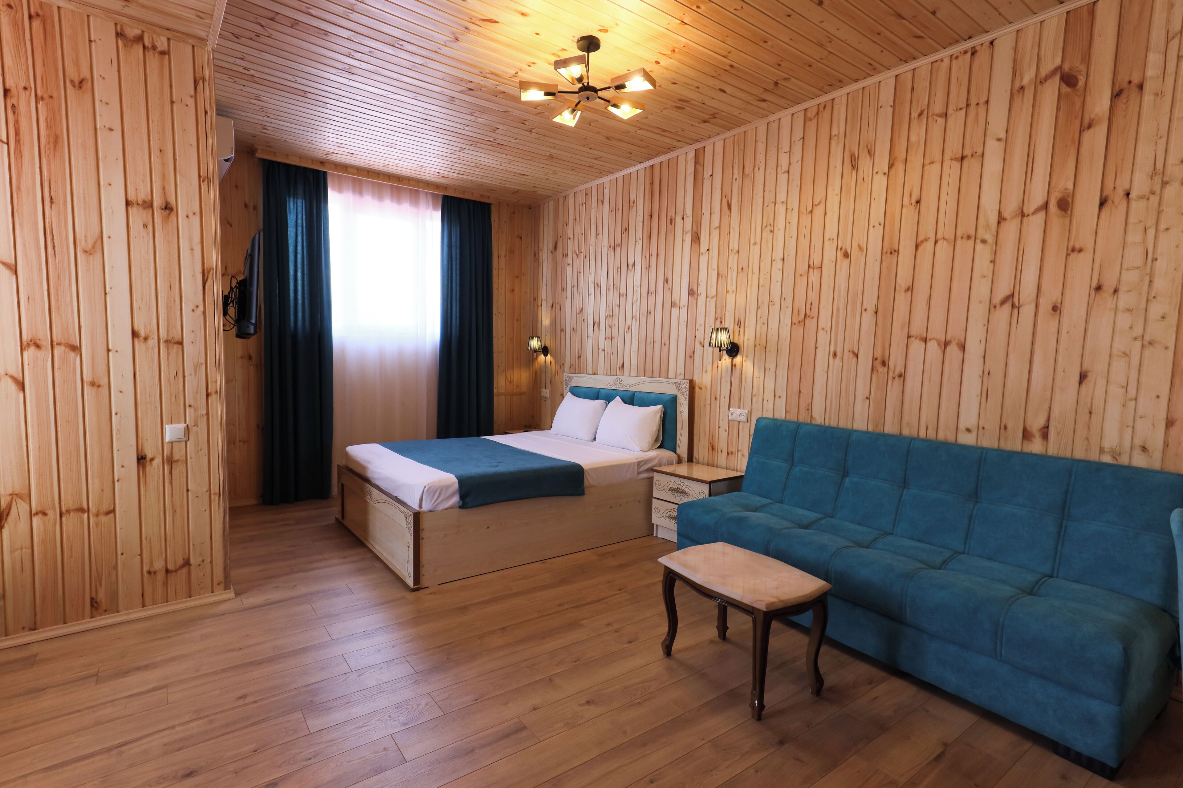 A wooden-paneled room with a bed, a blue sofa, a small table, and two wall-mounted lamps.