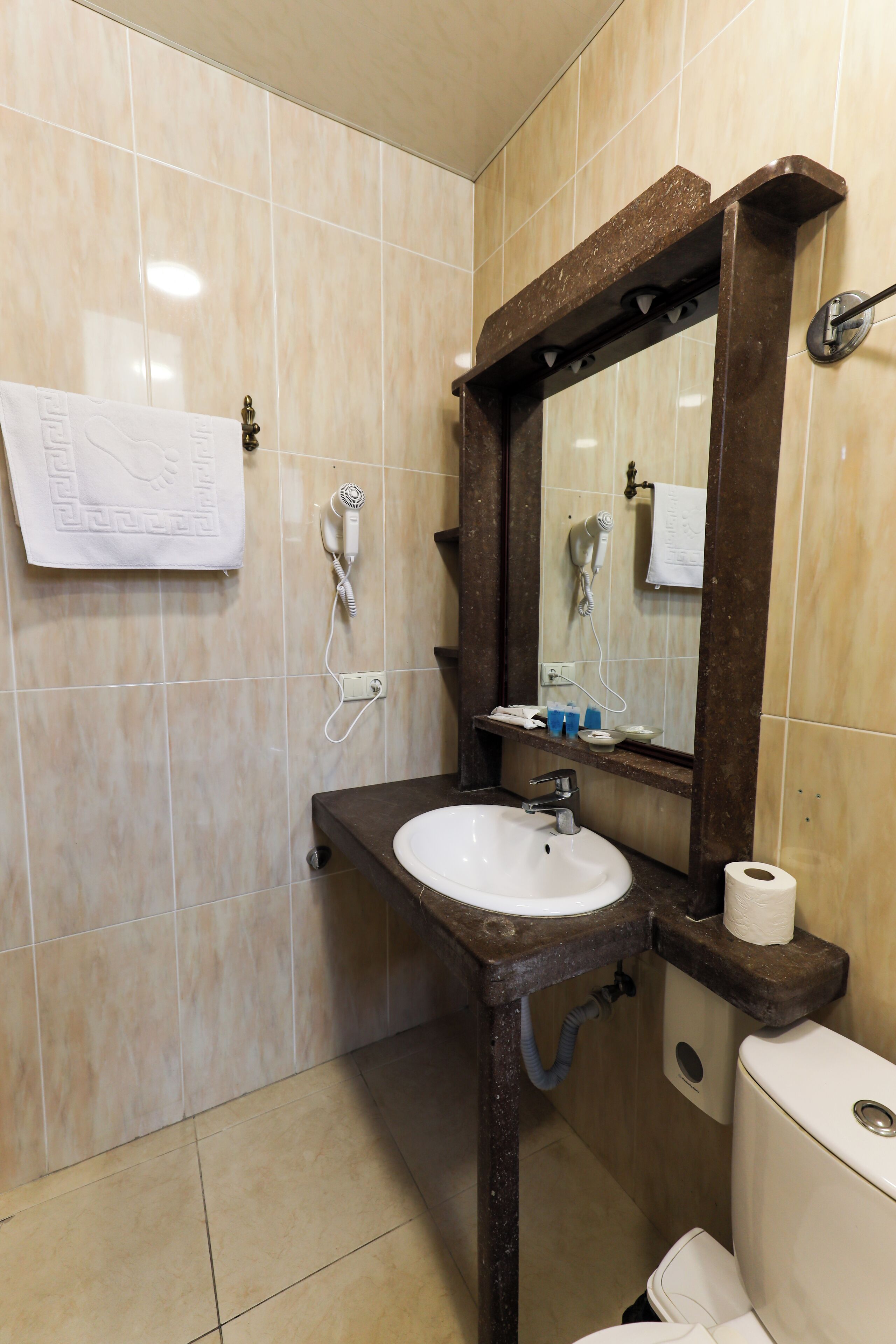 Ein Badezimmer mit einer Marmorfliesenwand, einem weißen Waschbecken, einem Holzkosmetikschrank, einem Spiegel, einem Handtuchhalter, einem Fön und einer Toilette.
