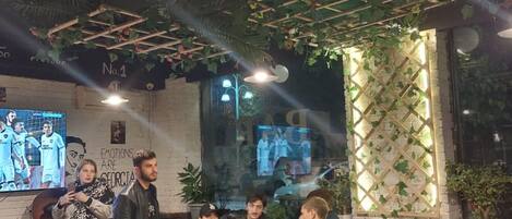 A cozy indoor seating area with tables and chairs, a TV screen displaying sports, and a decorative ceiling with plants.
