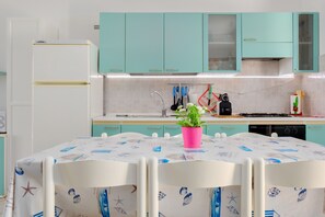 A kitchen with teal cabinets, a white refrigerator, a pink plant on the table, and a white tablecloth with blue patterns.